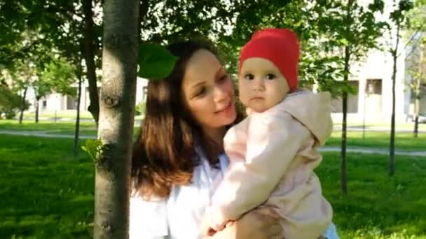 Feliz madre besa a su bebé en la mejilla y juega con una hoja en el parque de la ciudad al atardecer
 - Imágenes, Vídeo