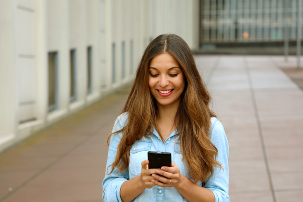 Beautiful young woman is using an app in her smartphone device to send a text message while standing in courtyard of office blocks - Foto, Imagen