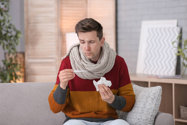 Beteg fiatal ember szenved a kanapén, otthon hideg hőmérővel - Fotó, kép