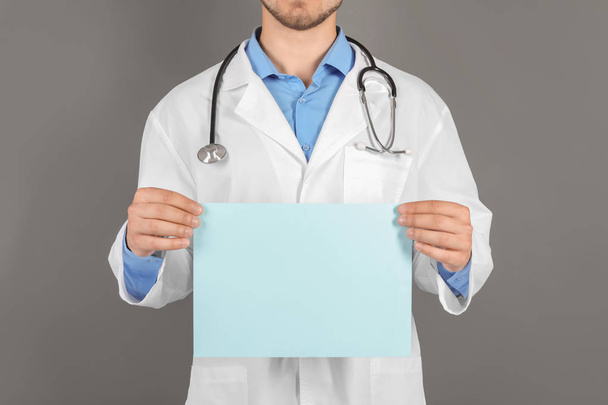 Male doctor holding blank sheet of paper on grey background - Foto, Bild