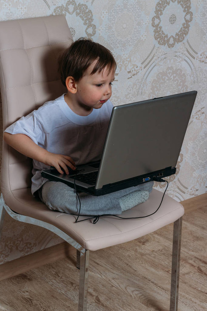 little boy in a white t-shirt sits on a chair, sofa with a laptop. looks into the camera. funny Face. concentration. - Fotó, kép