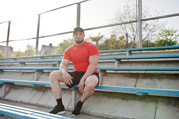Young brutal bearded muscular man wear on red shirt, shorts and cap at stadium. - 写真・画像