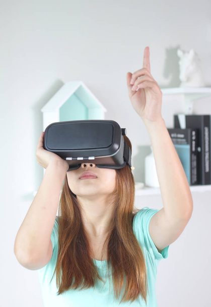 Νεαρή γυναίκα που φοράει γυαλιά εικονικής πραγματικότητας στο σπίτι. Ένα σετ κεφαλής εικονικής πραγματικότητας είναι ένα κεφάλι-mounted συσκευή που παρέχει εικονικής πραγματικότητας για τον κομιστή. - Φωτογραφία, εικόνα