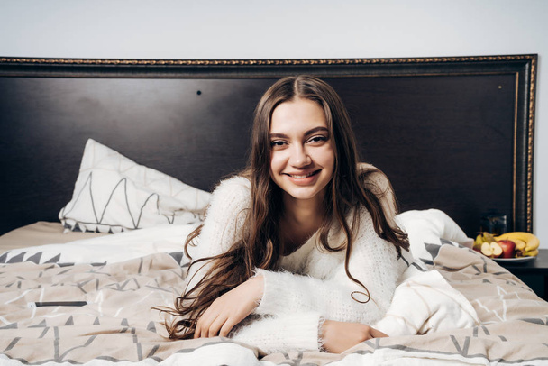 lovely long-haired girl is lying in bed late at night, resting after a work day and smiling - Фото, изображение