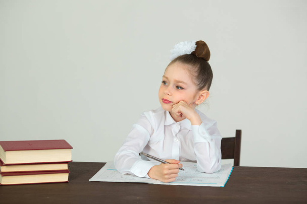 mooi meisje met een strik op haar hoofd zit aan een bureau met boeken op een witte achtergrond en huiswerk - Foto, afbeelding