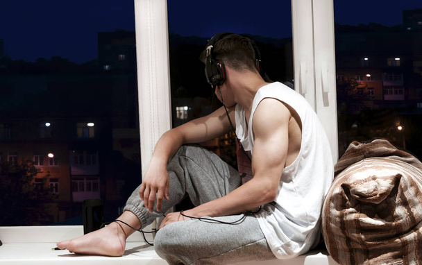stijlvolle jongeman, luisteren naar muziek, zittend op de vensterbank in de buurt van het venster - Foto, afbeelding