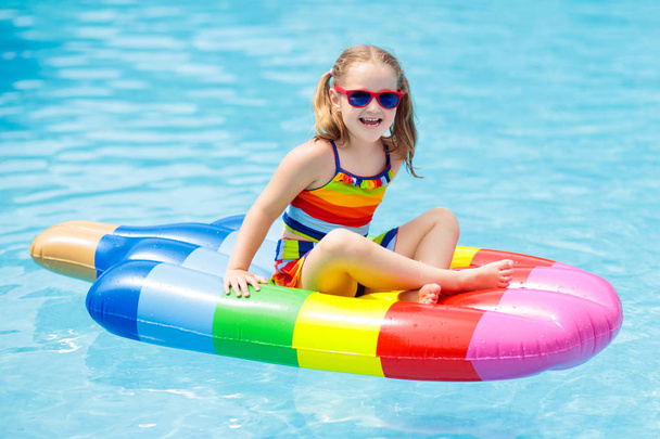 Happy child on inflatable ice cream float in outdoor swimming pool of tropical resort. Summer vacation with kids. Swim aids and wear for children. Water toys. Little girl floating on colorful raft. - 写真・画像