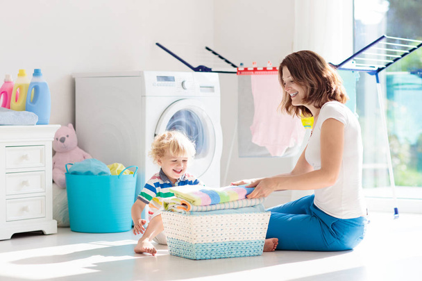Äiti ja lapset pesutuvassa pesukoneella tai kuivausrummulla. Perheaskareita. Nykyaikaiset kodinkoneet ja pesuaine valkoisessa aurinkoisessa kodissa. Puhdista pestyt vaatteet kuivaustelineessä
.  - Valokuva, kuva