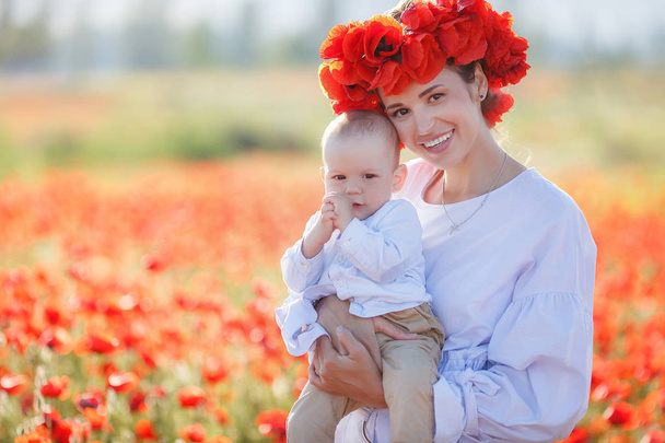 A beautiful young mother in a white long dress and a wreath of fresh red poppies with a small son in her arms enjoy the beauty of flowering red flowers, walking through a huge meadow on a sunny, summer day - Foto, Imagen