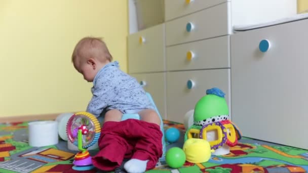 Smiling baby sitting on chamber pot with lots of toys and toilet paper around him in kids room - Кадры, видео