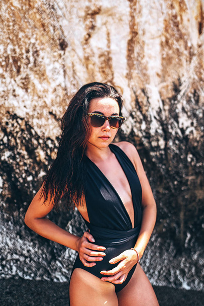 Brunette girl in black swimsuit posing near rock on the beach - Foto, Imagen