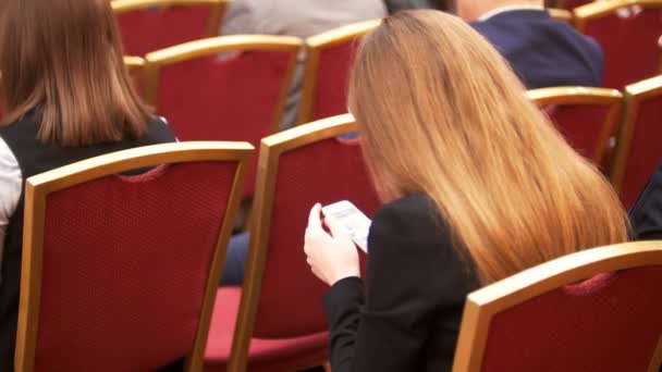 Ludzie siedzą w dużej sali na czerwone krzesła konferencji biznesowych, kobiety z smartphone w ręce - Materiał filmowy, wideo