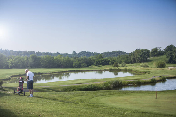 uomo maturo giocatore di golf con cappello vicino al suo golf cart su un campo da golf con lago in una giornata di sole
 - Foto, immagini