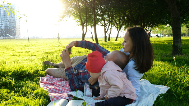 Happy family is laying on the pled and doing selfie with a baby at sunset in the park. Father takes pictures of themselves with the baby - Imágenes, Vídeo