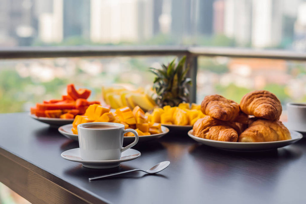 Breakfast table with coffee fruit and bread croisant on a balcony against the backdrop of the big city - 写真・画像