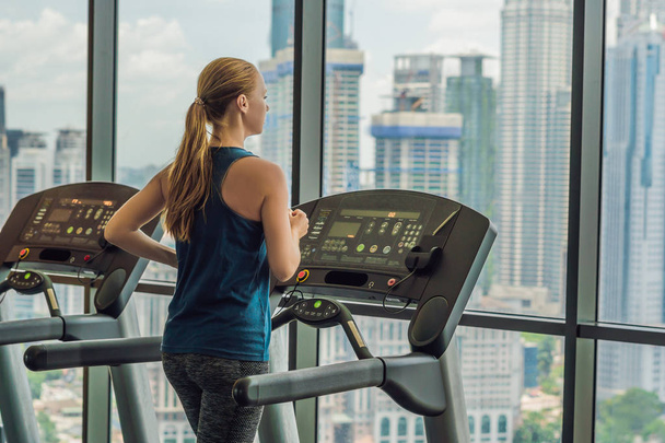 Beauty young woman exercising on treadmill in gym against the background of a big city. Sport, fitness, lifestyle, technology and people concept.  - Фото, изображение