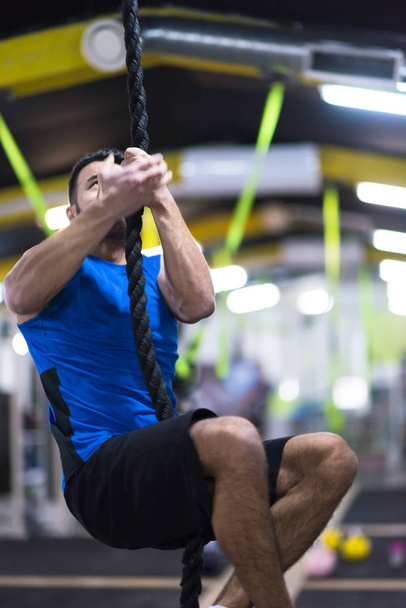 young muscular man doing rope climbing in cross fitness gym - Fotó, kép