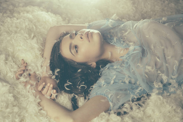 Dreamy girl lying on downy heap of white feathers. White fluff gently embracing young beautiful female in blue dress, bedtime fantasy concept - Фото, изображение