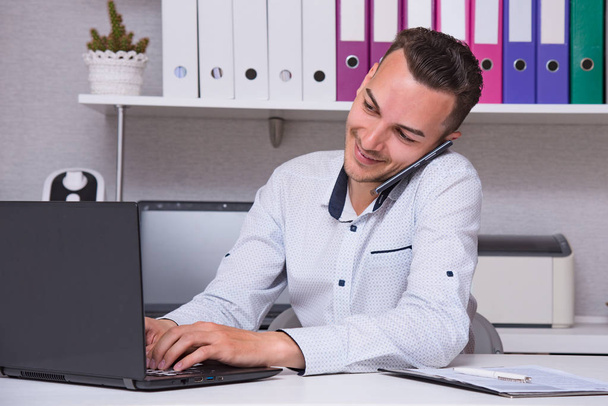 Portret przystojny młody mężczyzna Manager w biurze siedzi przy stole z laptopem rozmawia przez telefon. On siedzi tuż przed kamery, uśmiechając się i wyglądają poważnie - Zdjęcie, obraz