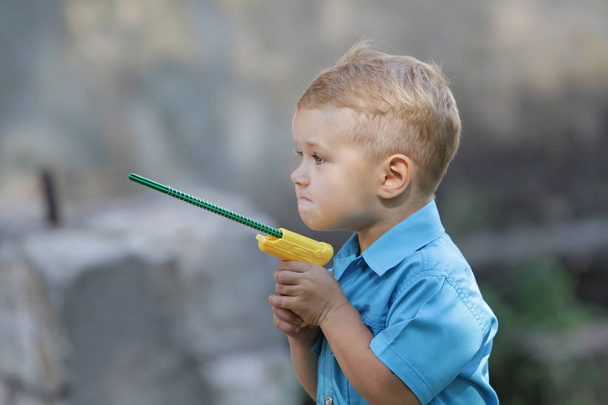 Ένα μικρό αγόρι παίζει με ένα παιχνίδι όπλο στην αυλή. Το παιδί παίζει και φαντασιώνεται παίρνει μια πλήρη ανάπτυξη και εκπαίδευση, εσωτερική ανάπτυξη. - Φωτογραφία, εικόνα