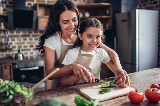 portrait of happy mother teaching her daughter to cut cucumber for the salad on the kitchen - Foto, Imagen