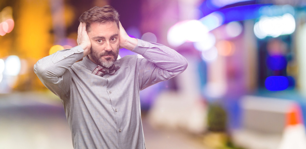 Średnim wieku człowiek, z brodą i muszka obejmujące uszy, ignorując przykry hałas, podłącza uszy aby uniknąć słyszenie dźwięku. Głośno muzyki jest problem. w klubie nocnym - Zdjęcie, obraz