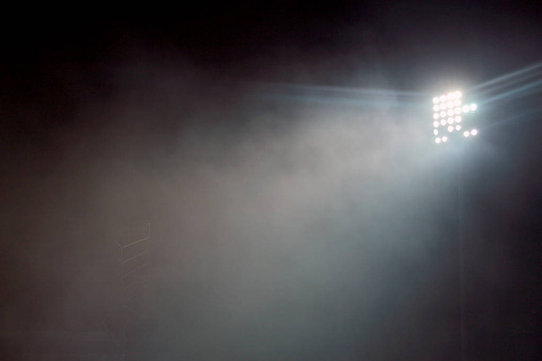 Light tower lit at a stadium during nightime. Stadium lights against dark night sky background. stadium lights and smoke. - Photo, Image