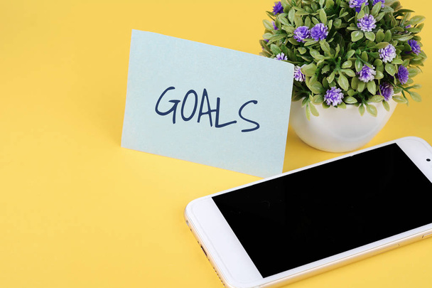 Paper note written with word "goals" and smartphone on yellow background. - Fotografie, Obrázek