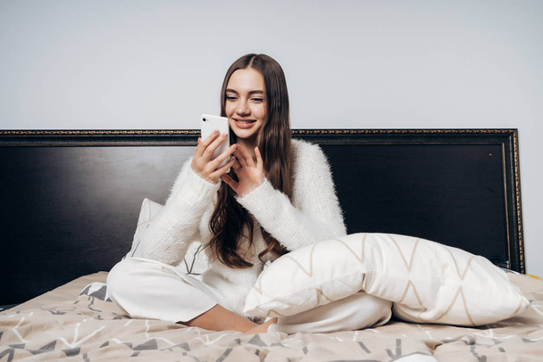 happy young girl wakes up early, sits in bed and looks into her smartphone, smiles - Фото, зображення