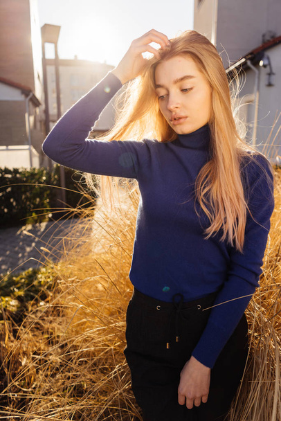 hermosa chica rubia con estilo en un suéter azul pasea por las calles de la ciudad, en los rayos del sol
 - Foto, Imagen