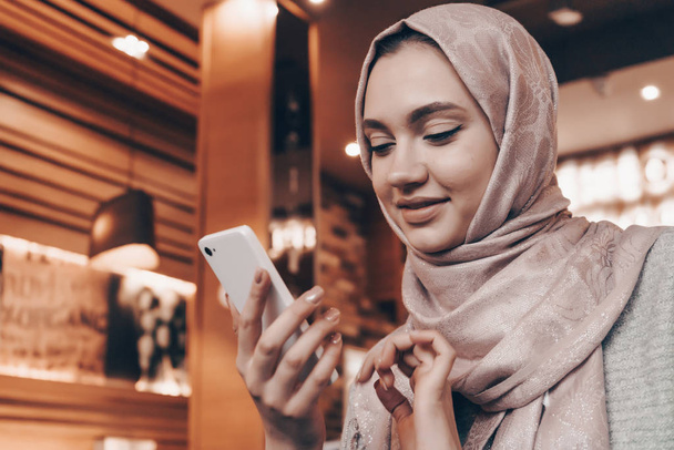 a beautiful Muslim girl with a headscarf on her head sits in a restaurant and waits for her food, writes a message on the smartphone - Foto, immagini