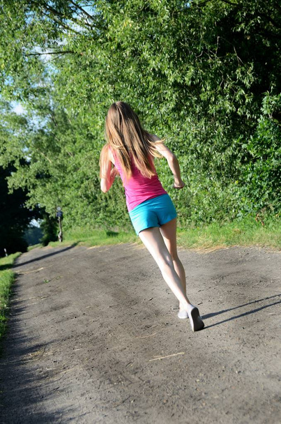 若い女性ランナー。女性は、緑の木々 に囲まれた未舗装の道路で実行されます。顔のない後ろからの写真. - 写真・画像