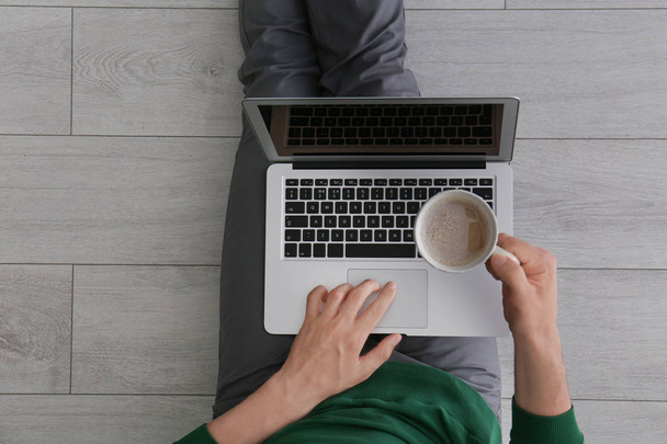 Jeune homme avec une tasse de café à l'aide d'un ordinateur portable assis sur le sol, vue sur le dessus
 - Photo, image