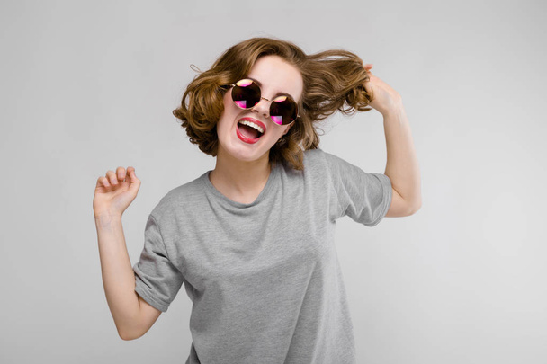 Charming young girl in a gray T-shirt on a gray background. Cheerful girl in round glasses holds a hand of hair - Photo, Image