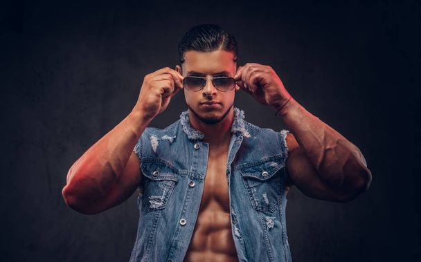 Sexy naked fashionable athletic man in a denim vest and sunglasses poses in a studio. Isolated on dark background. - Foto, Imagem