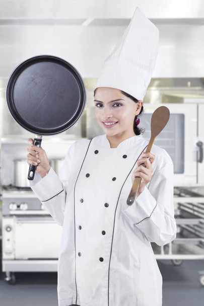 Portrait of an Indian female chef carrying spatula and frying pan while standing in the kitchen - Foto, Imagem