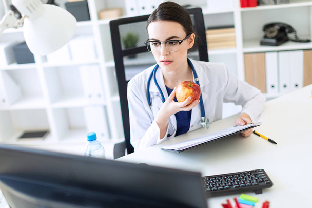 ein schönes junges Mädchen in weißem Gewand sitzt an einem Computertisch mit Dokumenten und einem Apfel in der Hand. - Foto, Bild