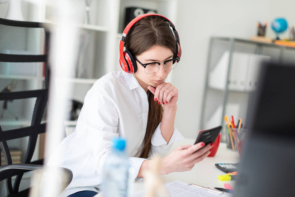 A young girl sits in headphones at a table in the office and holds a phone in her hand. - Foto, immagini