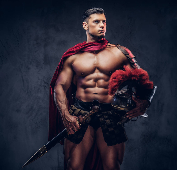 Brutaler alter griechischer Krieger mit muskulösem Körper in Kampfausrüstung posiert vor dunklem Hintergrund. - Foto, Bild