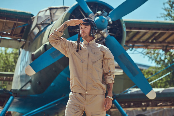 Dzielny przystojny pilot w pełnym biegu przekładni stojący w pobliżu retro samolotem wojskowym i wygląda odległości. - Zdjęcie, obraz