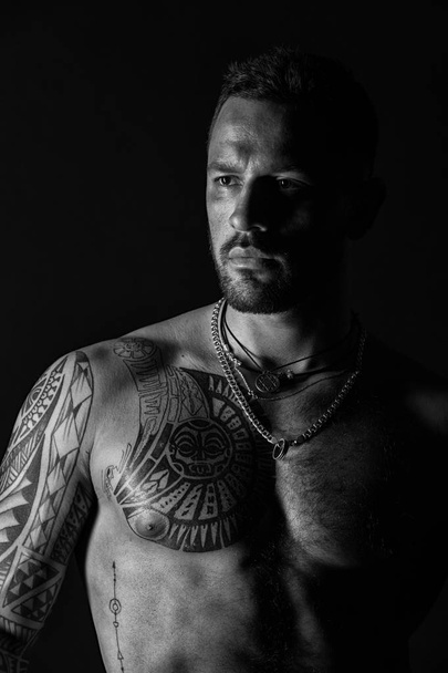 Bearded man with tattooed chest. Man with sexy muscular torso. Fit model with tattoo design on skin. Sportsman or athlete with stylish beard and hair. Bodycare or wellness and sport, black and white - Fotoğraf, Görsel