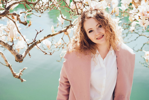 attractive stylish curly girl in pink coat posing in park in the sun and smiling - Фото, зображення