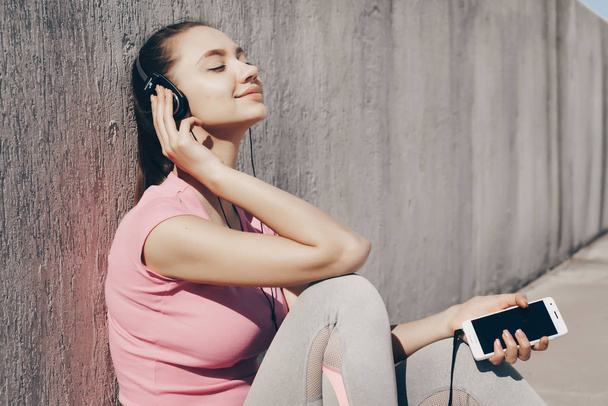peaceful young girl sitting on the ground in the sun, relaxing after a workout and listening to music on headphones - Fotó, kép