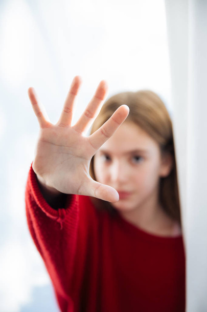 Sad cute little girl suffering depression and standing behind the window during a rainy day with raised hand  - Photo, Image