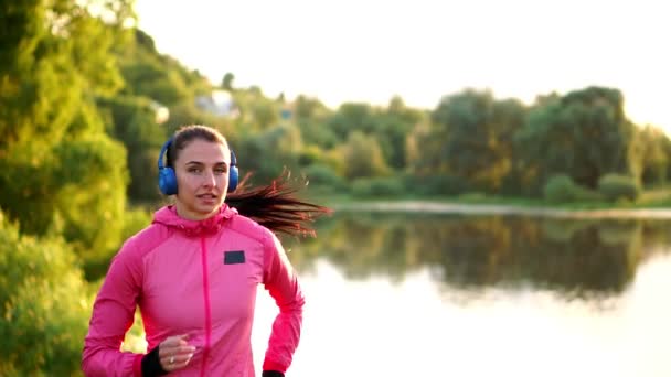 Brunette met lange haren in hoofdtelefoons loopt langs de rivier in het Park in de ochtend bij zonsopgang in de zomer in een roze jas en zwarte broek - Video