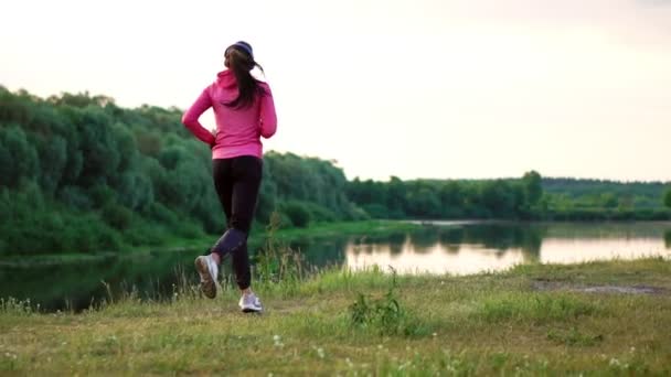 Een meisje in een roze jas en zwarte broek loopt in de buurt van de rivier in hoofdtelefoons voorbereiding voor de marathon - Video