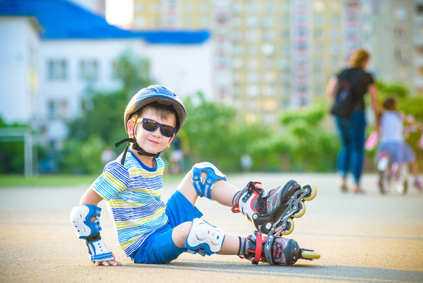 Happy boy in a protective helmet and protective pads for roller skating. Tired boy sitting and resting before start riding again. Active leisure summer holidays time for kids. - Photo, Image