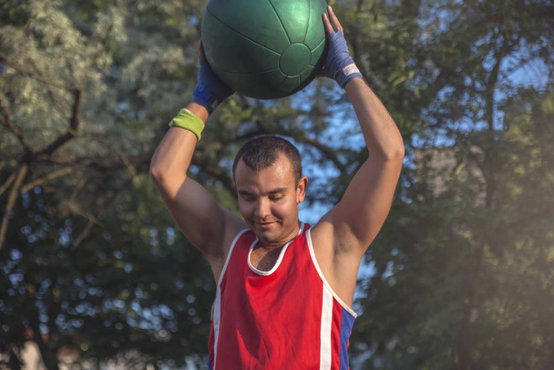 Νεαρός αθλητής εκτελεί αθλητικές ασκήσεις με μια μπάλα σε εξωτερικούς χώρους - Φωτογραφία, εικόνα
