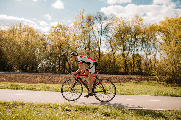 Велосипедист в шлеме и в спортивной одежде едет на велосипеде, вид сбоку. Тренировки на велосипедной дорожке, велоспорт
 - Фото, изображение