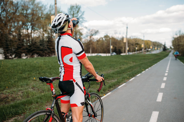Ciclista maschio in casco e abbigliamento sportivo beve acqua durante l'allenamento. Allenamento su pista ciclabile, ciclismo su strada asfaltata
 - Foto, immagini